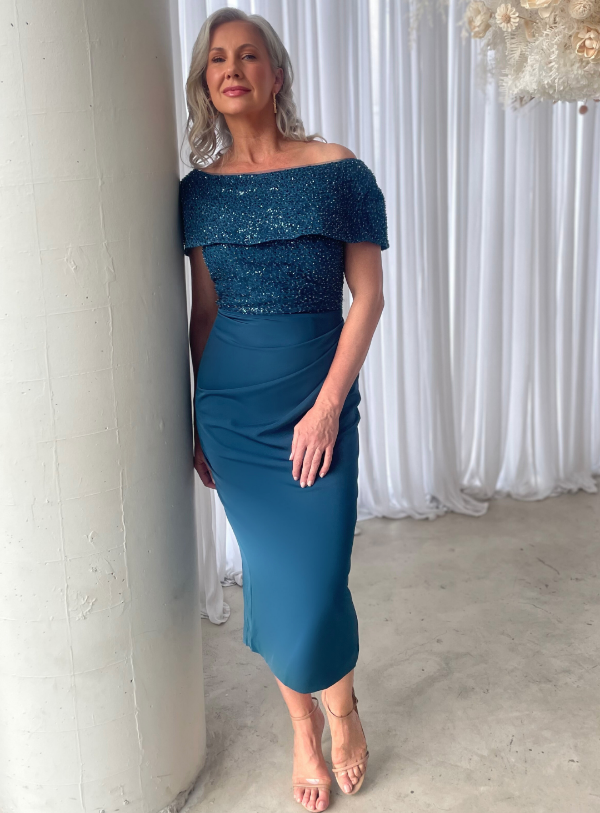 A woman wearing an off-the-shoulder mid-length blue dress with a beaded bodice and a woven satin crepe skirt.