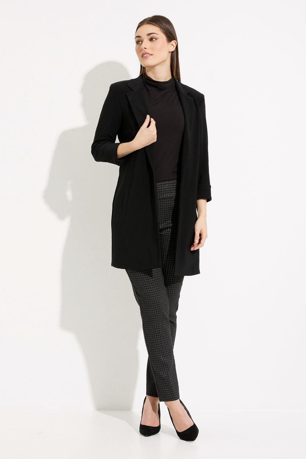 Woman wearing a black coat, black turtleneck, and gray pants on a white background