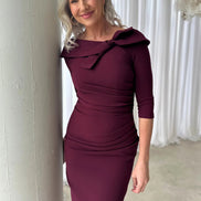 Woman wearing a burgundy dress with a large bow detail, standing against a white curtain.