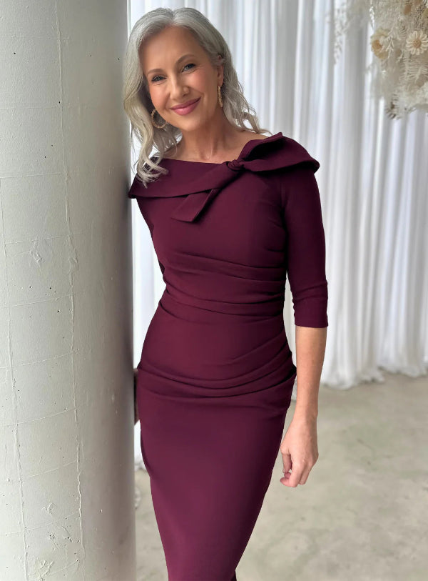 Woman wearing a burgundy dress with a large bow detail, standing against a white curtain.