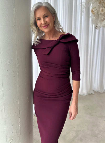 Woman wearing a burgundy dress with a large bow detail, standing against a white curtain.